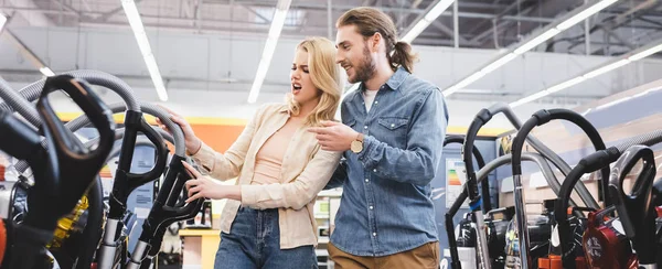 Plano panorámico de novio sonriente señalando con el dedo y mirando aspiradora con novia conmocionada en la tienda de electrodomésticos - foto de stock