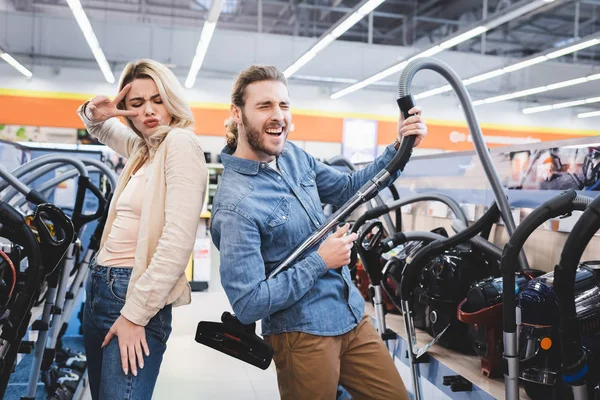 Boyfriend playing with vacuum cleaner and girlfriend showing peace sign in home appliance store — Stock Photo