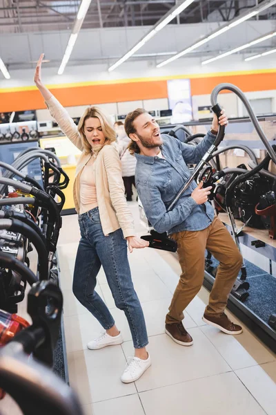 Novio jugando con aspiradora y novia bailando y cantando en la tienda de electrodomésticos - foto de stock