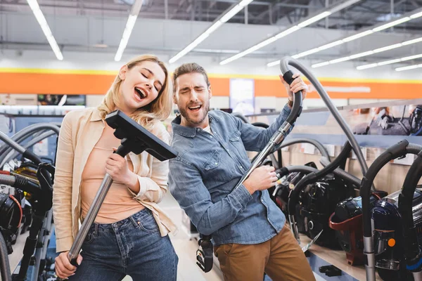 Boyfriend playing with vacuum cleaner and girlfriend singing in home appliance store — Stock Photo