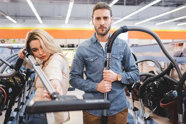 Serious boyfriend and girlfriend holding vacuum cleaners in home appliance store — Stock Photo