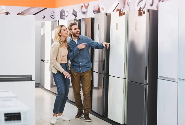 Smiling girlfriend and boyfriend pointing with fingers at fridge in home appliance store — Stock Photo