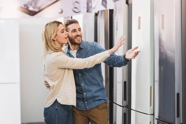 Smiling girlfriend and boyfriend pointing with hands at fridge in home appliance store — Stock Photo