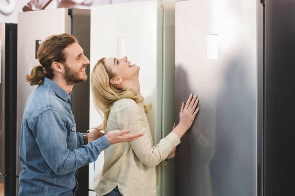 Side view of smiling boyfriend pointing with hand and girlfriend touching fridge in home appliance store — Stock Photo