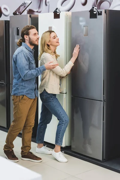 Boyfriend pointing with hand and smiling girlfriend touching fridge in home appliance store — Stock Photo