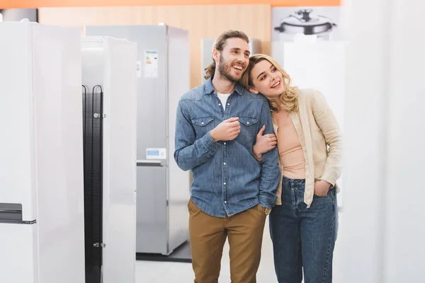 Novio y novia sonrientes mirando nevera en la tienda de electrodomésticos - foto de stock