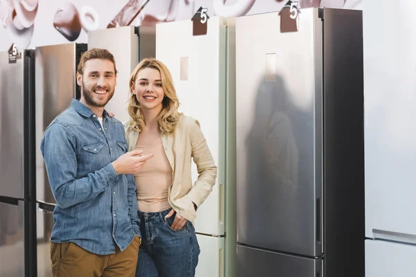 Smiling girlfriend and boyfriend pointing with finger at fridge in home appliance store — Stock Photo