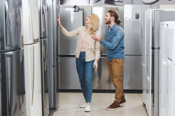 Smiling boyfriend pointing with hand and girlfriend touching fridge in home appliance store — Stock Photo