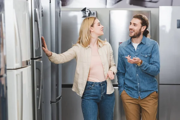 Fidanzato sorridente che punta con mano e fidanzata toccare frigorifero nel negozio di elettrodomestici — Foto stock