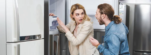 Панорамний знімок шокованого хлопця і дівчини, що стоїть біля холодильника в магазині побутової техніки — стокове фото