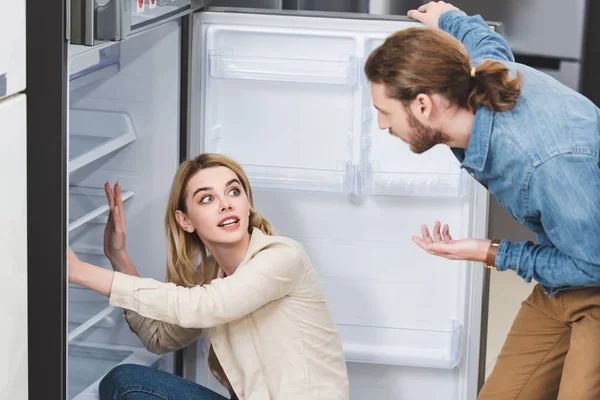 Novio hablando con impactada novia cerca de la nevera en la tienda de electrodomésticos — Stock Photo