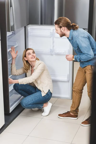 Namorado conversando com namorada sorridente perto de geladeira na loja de eletrodomésticos — Fotografia de Stock