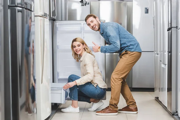 Sonriente novio mostrando como y novia sentado cerca de nuevo refrigerador en la tienda de electrodomésticos — Stock Photo