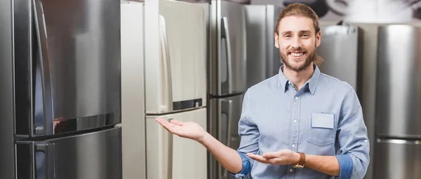 Prise de vue panoramique du consultant souriant pointant avec les mains au réfrigérateur dans le magasin d'appareils ménagers — Photo de stock