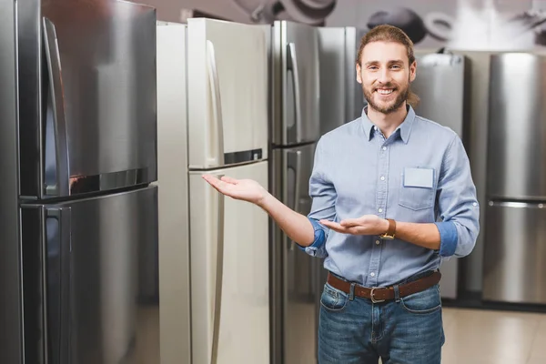 Smiling consultant pointing with hands at fridge in home appliance store — Stock Photo