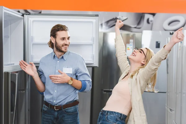 Consultant pointing with hands at fridge and smiling woman showing yes gesture in home appliance store — Stock Photo