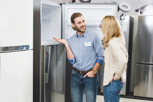 Smiling consultant pointing with hand at fridge and talking with woman in home appliance store — Stock Photo