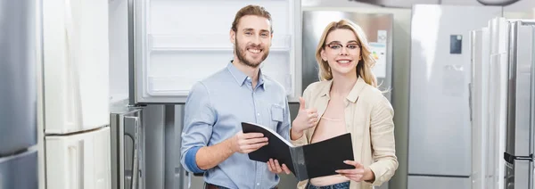 Panoramic shot of smiling consultant holding folder and woman showing like in home appliance store — Stock Photo