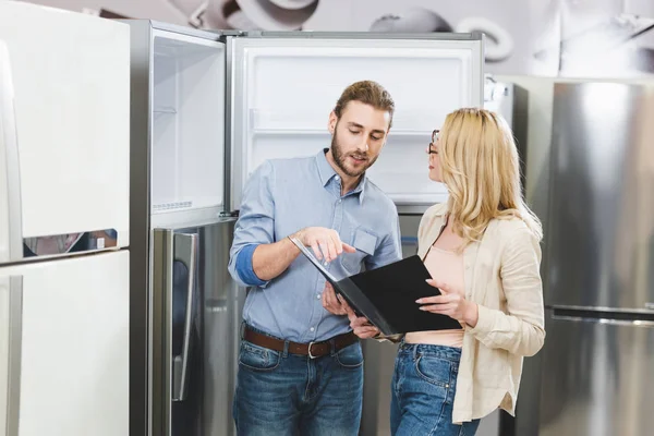 Consultant pointing with hand at folder and talking with woman near fridges in home appliance store — Stock Photo