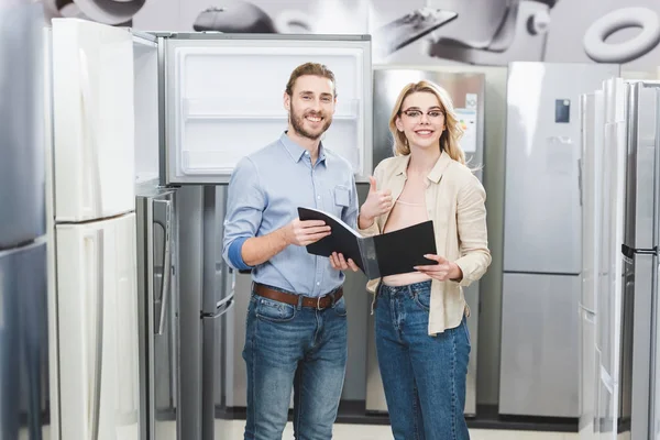 Smiling consultant holding folder and woman showing like in home appliance store — Stock Photo