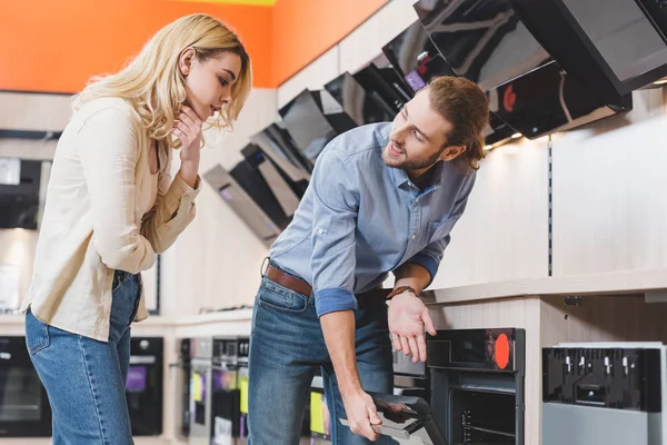 Consultor sonriente mostrando horno a mujer pensativa en la tienda de electrodomésticos - foto de stock