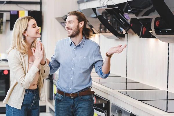 Smiling consultant showing shrug gesture and woman showing please gesture near cooker hoods in home appliance store — Stock Photo