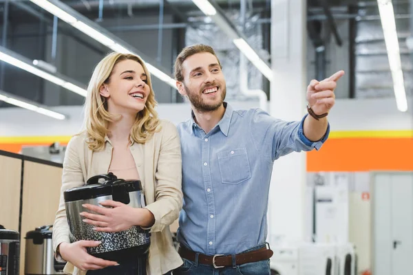 Consulente sorridente che punta con dito e donna tenendo cucina lenta nel negozio di elettrodomestici — Foto stock