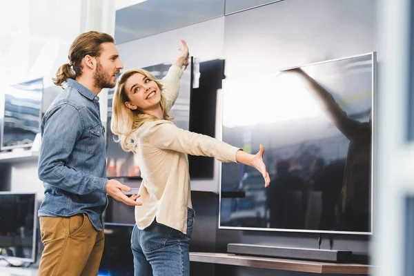 Sonriente novia apuntando con las manos en la televisión y mirando sorprendido novio en la tienda de electrodomésticos - foto de stock