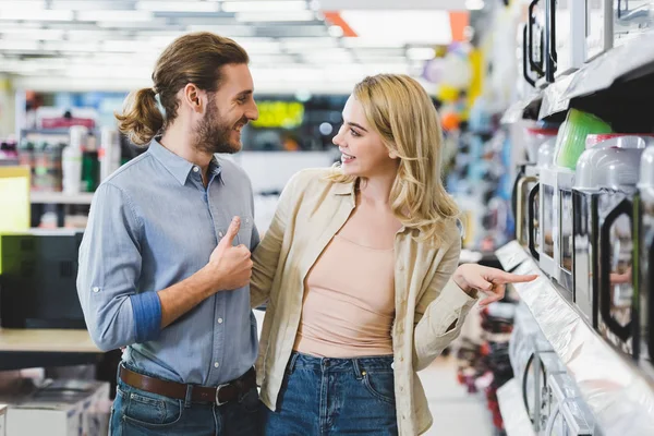 Consultor sonriente mostrando como y mujer señalando con el dedo en el microondas en la tienda de electrodomésticos - foto de stock
