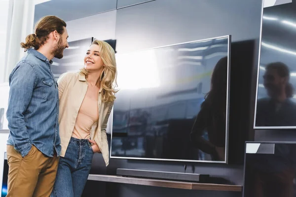 Sonriente novio y novia de pie cerca de la televisión en la tienda de electrodomésticos - foto de stock