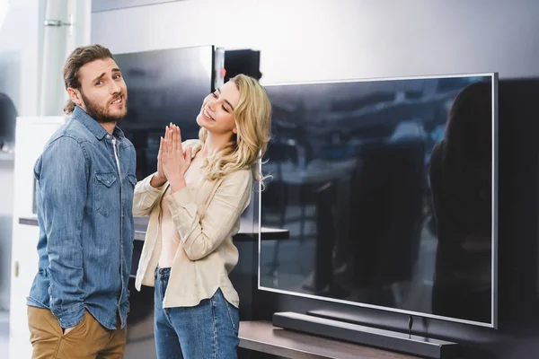 Confused boyfriend showing shrug gesture and smiling girlfriend showing please gesture near tv in home appliance store — Stock Photo