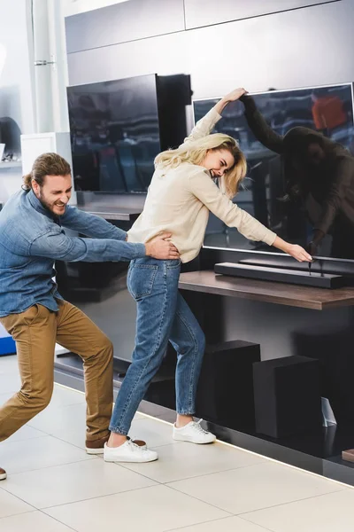 Angry boyfriend pulling girlfriend with tv in home appliance store — Stock Photo