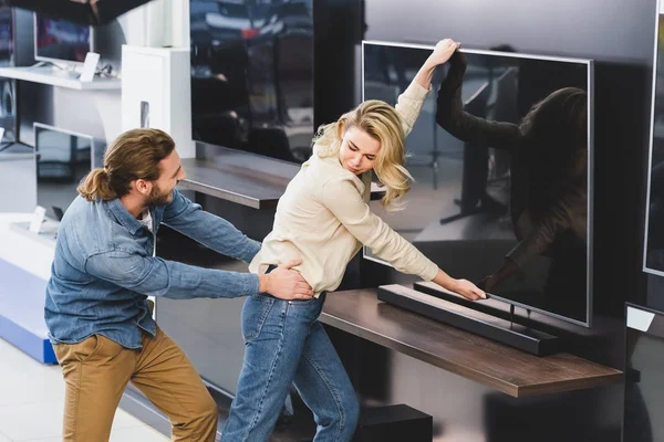 Novio enojado tirando de novia con televisión en la tienda de electrodomésticos - foto de stock