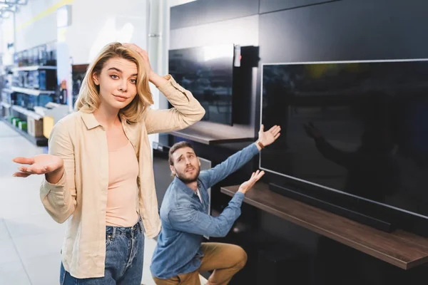 Messa a fuoco selettiva della donna confusa mostrando gesto scrollata di spalle e l'uomo che punta con le mani alla tv sullo sfondo in negozio di elettrodomestici — Foto stock