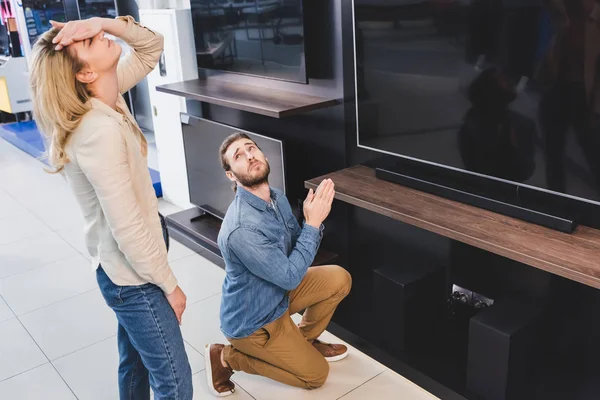 Хлопець показує, будь ласка, жест біля телевізора і втомилася дівчина торкається голови в магазині побутової техніки — стокове фото