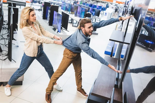 Freundin zieht traurigen Freund mit Fernseher in Haushaltsgerätegeschäft — Stockfoto