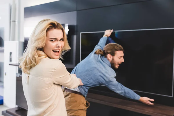 Selective focus of angry girlfriend pulling boyfriend with tv in home appliance store — Stock Photo
