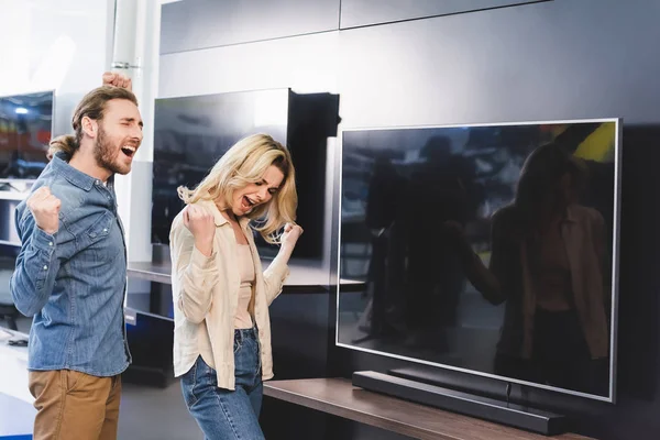 Feliz novio y novia mostrando sí gesto cerca de la televisión en la tienda de electrodomésticos - foto de stock