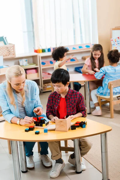 Bambino che gioca ai mattoni di un insegnante con bambini sullo sfondo nella scuola montessori — Foto stock