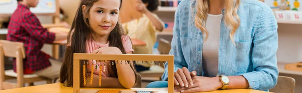 Selective focus of kid looking at camera while playing educational game by teacher in montessori school, panoramic shot — Stock Photo