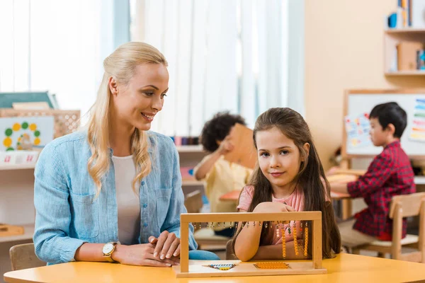 Lächelndes Kind, das in die Kamera schaut, während es ein Lernspiel des Lehrers in der Montessori-Schule spielt — Stockfoto