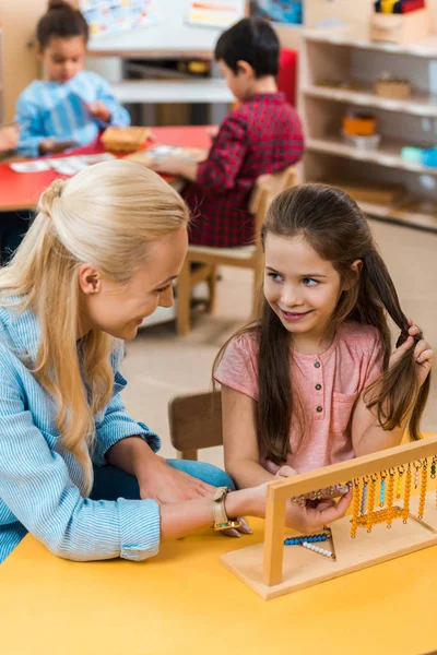Focus selettivo di bambini e insegnanti che si sorridono mentre giocano con i bambini in background nella scuola montessori — Foto stock