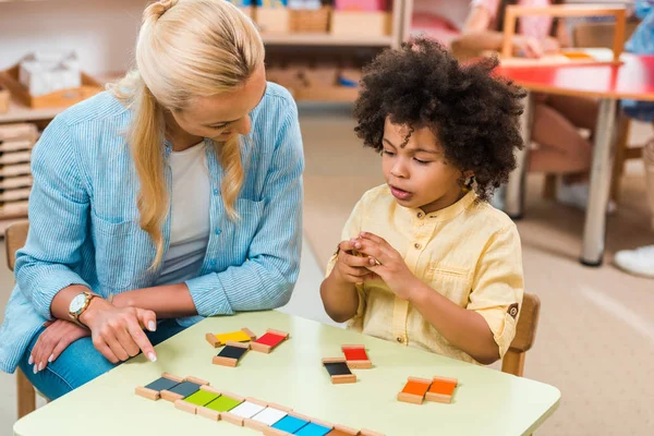 Niño afroamericano jugando juego educativo por maestro en la mesa en la clase montessori - foto de stock