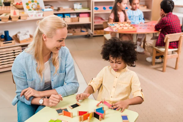 Selective focus of smiling teacher and child with wooden game at table and kids at background in montessori school — Stock Photo