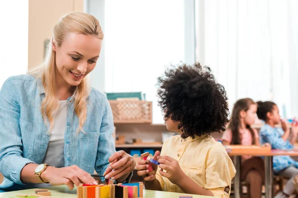 Foco seletivo de sorrir professor e criança jogando jogo educativo com as crianças em segundo plano na escola montessori — Fotografia de Stock