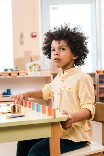 Pensativo niño afroamericano jugando juego de madera en la escuela montessori - foto de stock
