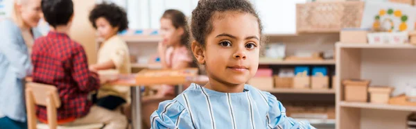 Focus selettivo del bambino afroamericano che guarda la macchina fotografica con insegnante e bambini sullo sfondo nella scuola montessori — Foto stock