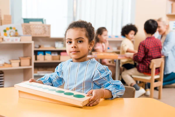Селективный фокус ребенка с деревянной игрой, смотрящего на камеру и учителя с детьми на заднем плане в школе Монтессори — стоковое фото