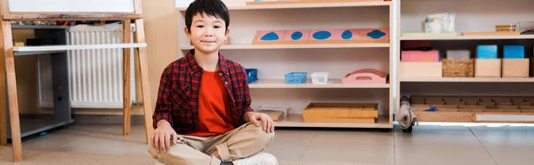 Colpo panoramico di sorridente ragazzo asiatico seduto sul pavimento nella scuola montessori — Foto stock
