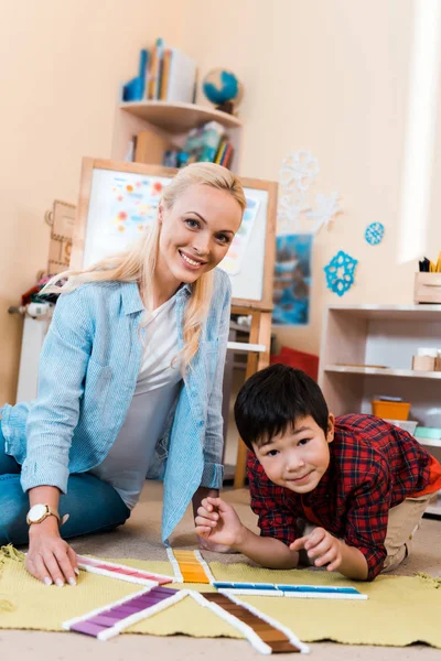 Selective focus of teacher and kid smiling at camera while playing game on floor in montessori school — Stock Photo